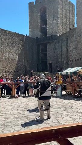 The video shows a sword fight between two individuals wearing armor. They are inside a stone courtyard, surrounded by a wooden barrier behind which a crowd of people stand watching. A large stone tower forms the backdrop, with more spectators on a balcony.  A green and white tent is visible on the right side of the frame. The sky is clear and blue. The armored figures clash swords, moving within the confines of the courtyard.