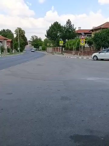 The video shows a street scene with houses and trees. There are road signs, including a speed limit sign of 30. A white car is parked on the right side of the road. The pavement is made of bricks. There are also some shops on the right side of the street. The sky is blue with some clouds. The video is taken from a moving vehicle, as the camera pans and shifts.