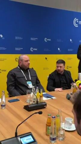Two men are seated at a long table with several microphones in front of them. The man on the left is wearing a black collared jacket and has a lanyard around his neck. The man on the right is wearing a black suit jacket. They are seated at a long wood table. There are several bottles of water and soft drinks on the table. A few sheets of paper and what looks like a cell phone are placed on the table. The background is split into a blue and yellow color. The yellow portion has several logos that contain the letters "MSC". Both men look to be in deep conversation.