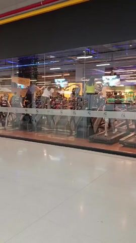 The video shows a gym from outside, seen through a glass wall. People are walking on treadmills inside the gym. The gym appears to be located inside a shopping mall or similar commercial building due to the white tile floor visible outside the glass wall and a yellow and red pipe running horizontally along the ceiling. There are multiple treadmills occupied with people exercising at different speeds. The gym's name "Fit" is displayed on the glass wall. The lighting inside the gym is bright, with several monitors visible above the treadmills.