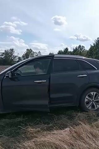 The video shows a dark-colored SUV stopped on the side of a dirt road in a rural area. The front passenger door of the SUV is open, and a person can be seen sitting in the driver's seat. The car is partially in a field of dry grass and dirt, with trees and vegetation visible in the background under a partly cloudy sky. The camera pans across the landscape, showing more of the field and the sky. The person in the car appears to be looking around, possibly at the surrounding environment or the camera.