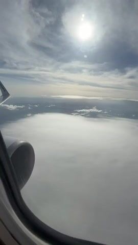 The video presents a view from an airplane window, showcasing a bright sun partially obscured by wispy clouds in a blue sky. Below, a thick layer of white clouds stretches out to the horizon, creating a sea of white. The edge of the plane's engine is visible in the lower left corner of the frame, adding a point of reference. The sunlight reflects off the cloud layer, creating a bright, diffused light across the scene.