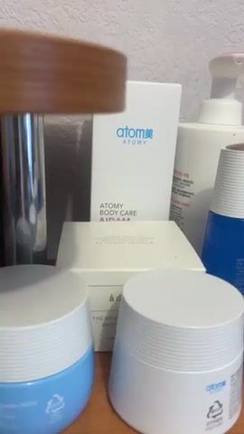 The video displays a collection of skincare products, primarily from the brand "Atomy". The products are arranged on a wooden surface against a textured wall. The camera pans across the collection, showing various bottles, tubes, and jars. Visible products include an "Atomy Body Care" lotion, an "Essence Sun" sunscreen, and an "Eye Cream". Several other containers with "Atomy" branding are also present, some with Korean text. The lighting is neutral, and the focus is on the products themselves. There are no people visible in the video.