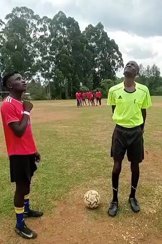The scene shows a soccer field with a referee in a bright yellow shirt and black shorts, black socks, and black cleats standing near a soccer ball. Two soccer players, one in a red jersey with black shorts and the other in a blue and yellow uniform with an orange armband, are standing next to him. In the background, a group of soccer players is standing together. The referee points to the ground and speaks. The video appears to show the officials explaining the rules of the game to the players. The field appears to be a grass field that is partly bare. Trees are visible in the background. The weather appears to be overcast.