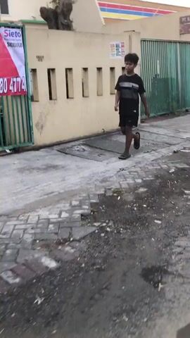 A young boy walks on a sidewalk in front of a building. He is wearing a black t-shirt and black shorts, and he is walking from right to left in the frame. The boy appears to be walking with a relaxed gait. He is wearing sandals. The sidewalk is made of concrete and cobblestone. There is a drain cover on the sidewalk. The building behind the boy is light yellow and has several vertical slits in its facade. A green metal fence runs along the front of the building. A banner with red and white text and a picture of a house is attached to the fence. A building with a colorful striped awning is visible in the background. The ground is wet in places, suggesting recent rain. The lighting suggests it is daytime, possibly late afternoon. The scene appears to be an urban or suburban environment in Ngaban, East Java. There is no indication of the boy's emotional state. Text visible on the banner reads: "Sieto Real Estate Agent SIDDARJO" and "0800 4774".