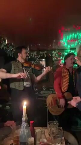 Two musicians are performing in a dimly lit room. One musician on the left is playing the violin and wearing a green vest. The other musician is playing the guitar and wearing a red jacket. They are both singing. There is a candle on the table in front of them, and several glasses and bottles. There are Christmas lights in the background. A person wearing a blue sweater stands to the right of the musician playing the guitar.