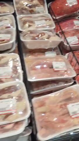 The video shows a close-up view of packaged raw meat products displayed in a retail store refrigerator. The products are primarily ground meat and what appears to be chicken or pork pieces. They are arranged in clear plastic packaging on white styrofoam trays, stacked in tiered metal shelving. The lighting is artificial and appears to be fluorescent, typical of a grocery store environment. There are no people visible in the footage. The focus remains on the products, with some labels visible but not clearly legible. The camera angle is slightly angled downwards, providing a view of the product arrangement. The overall impression is that of a refrigerated meat section in a supermarket.