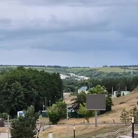A drone flies over a landscape with trees and hills. A cloudy sky looms overhead. Several birds are seen flying in the distance.