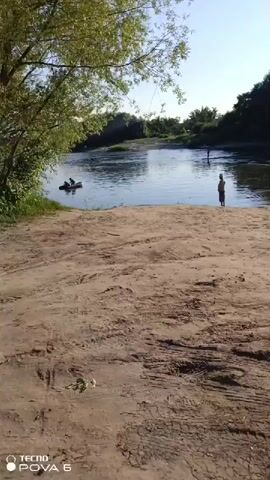 This video depicts a serene natural scene by a river. The foreground is a sandy beach with tire tracks, suggesting recent vehicle activity. In the middle ground, a calm river flows, bordered by lush green vegetation and trees on the opposite bank. The water reflects the clear, bright sky.

In the distance, two individuals are visible in the river. One is standing in the water, appearing to be fishing or enjoying the coolness. Another person is further back, also in the water. The overall atmosphere is peaceful and tranquil, indicative of a warm, sunny day, likely late afternoon given the soft lighting and long shadows. The presence of trees suggests a rural or natural setting, consistent with a location outside a major city. The background is dominated by a dense line of trees, creating a sense of depth and a natural barrier.

The visible text at the bottom left corner of the screen reads "TECNO POVA 5," which is likely the model of the mobile device used to record the video. No other text is present. The emotional tone evoked by the scene is one of relaxation and simple enjoyment of nature.