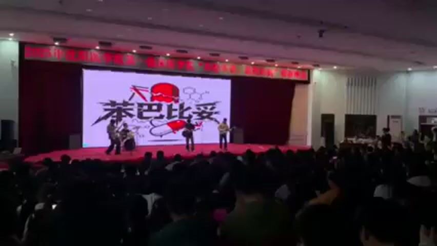 A wide angle view of a live band performing in front of a large audience. The band consists of drums, guitar, bass, and a lead singer. They are on a stage with a large screen behind them. The screen displays a logo, which contains text and a drawing, which includes a pill and a molecular diagram. The audience is seated and watching the performance. The room appears to be a large auditorium.