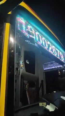 The video shows the front and interior of a yellow and red truck at night. The truck has bright LED headlights and an illuminated digital display above the windshield. The interior of the truck is visible through the windshield, including the steering wheel, dashboard, and seats. The video also shows a close-up of the truck's headlights, which are brightly illuminated and feature a modern design with multiple LED elements. The environment appears to be an outdoor setting, likely a street or parking lot, given the presence of dark shadows and what appears to be asphalt on the ground.