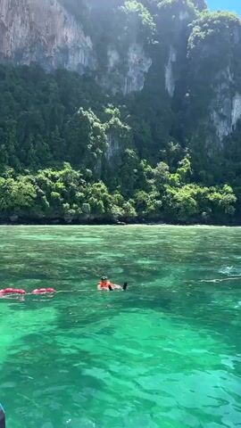 The video shows a person in the water wearing an orange life vest and snorkeling gear. The water is clear and turquoise, with sunlight filtering through it. In the background, a lush green, rocky cliff face rises from the water, covered in trees. There are red buoys in the water to the left of the person. The person is floating on their back, appearing relaxed. The scene suggests a tropical vacation or recreational activity in a beautiful natural setting.