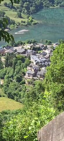 The video provides a high-angle shot looking down a steep, overgrown hillside towards a town situated beside a body of water. In the foreground, weathered, abandoned train tracks are visible, partially covered by vegetation, running alongside a concrete structure. Further down the slope, a long, narrow building with a flat roof is situated in a grassy area, with a series of pipes or cables running parallel to it. Beyond this, a cluster of buildings, characteristic of a village or small town, can be seen. The town is nestled on the edge of a tranquil, blue body of water, with rolling green hills and trees forming the backdrop. The overall impression is one of a scenic, somewhat remote location, with elements of past industrial activity now being reclaimed by nature.