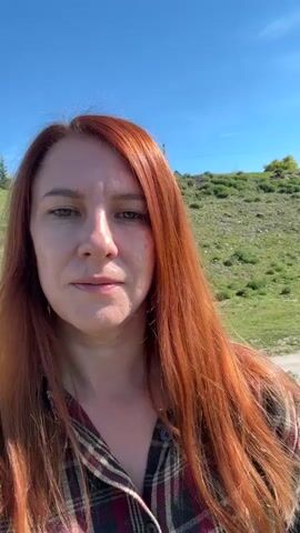 A woman with long red hair is filming herself, facing the camera. She's wearing a plaid shirt, and the background changes as she pivots around a river. In the background is a bright blue sky with a few puffy clouds, a wide river with forested banks, and mountains in the distance.