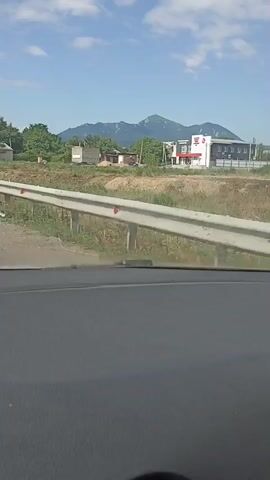 The video shows a view from inside a vehicle, looking out at a rural landscape. The foreground is dominated by the dark interior of the car, specifically the dashboard and windshield frame. A white guardrail with red reflectors runs horizontally across the middle ground, separating the road from a grassy embankment. Beyond the embankment, there are several buildings and trees. In the distance, a mountain range under a blue sky with scattered clouds is visible. The footage appears to be captured while the vehicle is in motion, as suggested by the slight blur and the changing perspective. The time stamps indicate the video lasts for approximately 4 seconds.