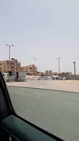 The video shows a scene from a moving car, looking out the driver's side window. The camera pans slightly, showing a street with small, white, enclosed booths along one side. In the background, there are several multi-story buildings in a light brown color, with visible windows. A truck with people seated under a canvas cover passes by. In the later part of the video, a young person, a woman in a black robe, and a man are seen walking on the side of the road. The sky is bright and clear, suggesting a sunny day. The road appears to be in a relatively quiet area with buildings in the background.