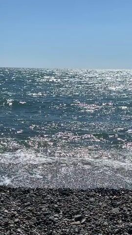 The video shows a close-up of a shoreline with dark grey pebbles in the foreground. The sea stretches out into the distance, reflecting the bright sunlight, creating a shimmering effect on the water's surface. Gentle waves are breaking on the shore, creating white foam. The sky above is a clear, bright blue. There are no people or objects visible in the video.
