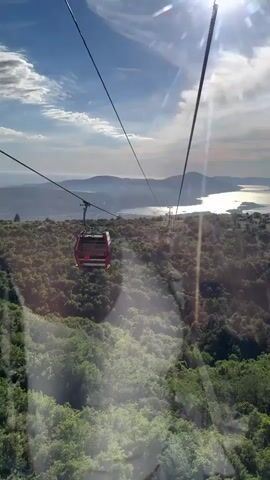A cable car descends a mountain, traveling over a forest with a body of water and hills in the background. The sun is shining brightly in the upper right corner of the frame, creating lens flare. A red cable car is visible in the lower left portion of the frame, and another cable car is further in the distance. The video is shot from the perspective of a person inside a cable car.