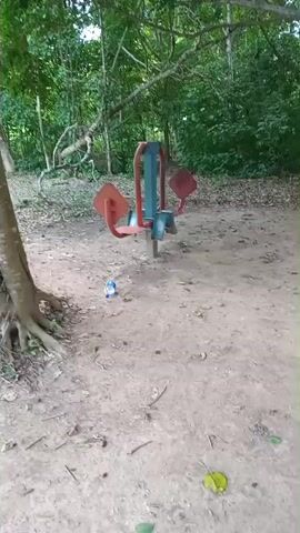 A person is walking on a dirt path in a wooded area. There is some outdoor exercise equipment visible, including what appears to be a pull-up bar setup and some other red and blue machines. A basketball court with colored sections is also visible in the background. The person is wearing a red t-shirt and blue shorts. They stop and pose for the camera, giving a thumbs up. The video is shaky and the focus is not always clear.