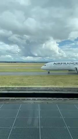 From behind a window, a static shot shows an Air France airplane on the tarmac. The plane is white with the company's name and logo in blue and red. Behind the plane is a strip of green grass that blends into the grey tarmac of the runway. The sky is partially overcast with large, billowing clouds. Below the window is a metallic, grid-like structure.