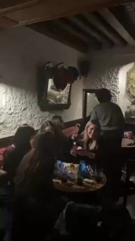 The video depicts a crowded pub interior. Several people are seated around a table cluttered with drinks. A staff member, wearing an apron, moves among them. The camera pans across the space, showing a bar area filled with people standing and talking. Decorations, including lanterns and lights, hang from the ceiling. The pub has a rustic feel, with exposed wooden beams and stone walls. An old clock hangs on a pillar.