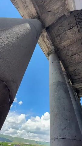 The video pans upwards to show the underside of a stone roof supported by large stone columns. The roof has intricate geometric carvings. Beyond the columns, the sky is a bright blue with scattered white clouds. In the distance, green hills are visible. The bottom of the video shows a group of people gathered near the base of the columns, looking up at the structure. Some are holding cell phones, presumably to take pictures. The overall scene suggests a visit to a historical site, possibly an ancient temple or ruin.