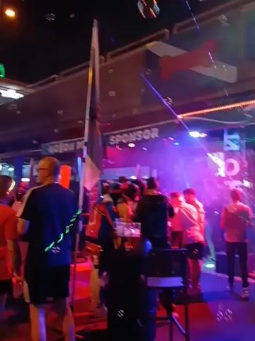 The video shows a nighttime outdoor event with many people gathered and dancing. Bubbles float in the air, and there are signs indicating sponsors. Some individuals are wearing minimal clothing, including harnesses and underwear. People are interacting and moving around, some dancing and others talking. The environment includes buildings, lights, and what appears to be a stage area.