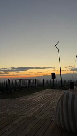 The media file captures a serene sunset scene at an outdoor venue, likely an event space or a lounge area, in Jrvej, Armenia. The setting features a wooden deck with a striped beanbag chair in the foreground, suggesting a relaxed atmosphere. Beyond the deck, a metal railing separates the area from a vista of rolling hills and a distant city skyline, illuminated by the fading sunlight.

The sky is a canvas of soft gradients, transitioning from a pale yellow near the horizon to a dusky grey overhead. Wispy clouds are scattered across the sky, catching the warm hues of the setting sun. A prominent object in the scene is a tall, black stand with an angled fixture at the top, possibly for lighting or a camera. Near the railing, a black speaker stands on a tripod, hinting at a potential event involving music or a presentation. To the right, a large, inflatable screen is partially visible, suggesting that an outdoor movie screening or a similar event might be taking place. The time of day is clearly sunset, indicated by the low sun on the horizon and the soft, warm light. The weather appears to be clear and pleasant, conducive to an outdoor gathering.

Although no specific activity is explicitly shown in progress, the setup with the screen and speaker implies an organized event. The overall mood conveyed is tranquil and picturesque, with the natural beauty of the sunset complemented by the prepared venue. The location cues of the hilly landscape and city lights below, combined with the given location context, firmly place the scene in Jrvej, Armenia. No visible text is present in the media file.