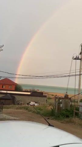 The video captures a rural scene under a cloudy sky, with a prominent rainbow arcing across the frame. In the foreground, a dirt path leads towards the horizon. To the left, a rustic building with an orange roof is visible, partially obscured by a smaller structure and some hay bales. Beyond these buildings, a body of water, likely the sea, stretches towards the horizon. The middle ground is dominated by utility poles and overhead wires, some appearing weathered and tangled. One pole supports a large electrical transformer. Dense green foliage and trees fill the right and bottom portions of the frame, creating a natural backdrop. The overall impression is one of a tranquil, possibly post-rainy day landscape.