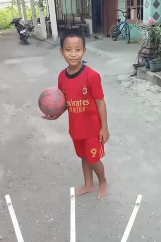 A young boy, barefoot, wearing a red t-shirt and shorts with the number 9 on the shorts, stands outdoors holding a red basketball. He is smiling and looking towards the camera. In front of him is a white, circular object with multiple white spokes extending outwards, possibly a drying rack. Behind him, there is a dirt or gravel ground, a building with windows and a door, a motorcycle, and a bicycle. The boy appears to be in a residential area. The timestamp indicates the video is only a few seconds long.