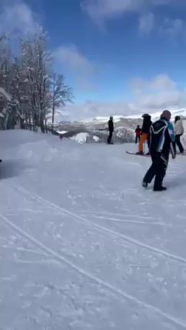 The video shows a snowy landscape on a bright day, likely at a ski resort. There are bare trees with some snow clinging to the branches on the left side of the frame.  Several people are standing and skiing on a snow-covered slope, most visible in the right half of the video. A snowmobile, ridden by someone wearing a green jacket, is on the left side of the frame, moving slowly away from the camera along a track in the snow. In the background, there is a view of snow-covered hills under a blue sky dotted with clouds.