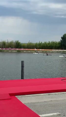 The video shows a drone view of a kayaking race on a calm lake. Several kayakers are visible in the distance, paddling in a line. The banks of the lake are lined with trees and flags. A red carpet is visible in the foreground, suggesting a formal event. The sky is cloudy, and the water appears to be dark. There are a few buoys marking the course. The scene is quiet and peaceful.