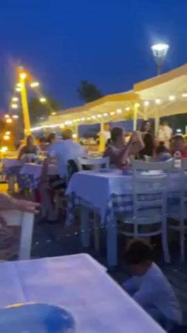 The video shows a night time scene at an outdoor beach restaurant, with a focus on a few people seated at tables. The area is illuminated by strings of lights hanging overhead. In the foreground, there are empty plates and cutlery on a table covered with a white tablecloth. The camera pans across, revealing two young men seated at separate tables. The first is wearing a multi-colored sports jersey, while the second is in a black shirt and smoking a cigarette. The sea can be seen in the background, under a deep blue sky. There are other people dining at nearby tables. The atmosphere is relaxed and social.