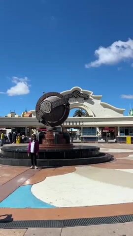 The video shows the entrance to Parque Warner Madrid, with a woman standing in front of the Warner Bros. globe fountain, and someone taking her picture with a camera. The fountain is centered in a colorful, patterned plaza. The architecture behind the fountain is a wide, arched building, typical of theme park entrances, with food vendors. The sky is blue with a few scattered clouds. The video has background music.