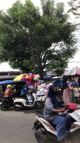A street market scene. A large tree with green foliage dominates the upper portion of the frame, with power lines running through it. Below the tree, a market stall is set up. Several colorful balloons are displayed, some tied together and some in bunches, attached to poles. There are also bags of merchandise on display. In the foreground, a paved area is visible. Two motorbikes are present. One is black, with a person wearing a yellow helmet and an orange and brown jacket on it, facing away from the camera. The other is white, with a person wearing a blue hijab and a light blue top on it, also facing away from the camera, with bags on the front of the motorbike. Another motorbike is partially visible to the left. People are moving around the stalls. The overall impression is of a busy outdoor marketplace.