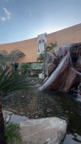 The video shows an artificial waterfall cascading into a pond. The waterfall is constructed from large, irregularly shaped reddish-brown and gray rocks. The pond is dark with some foam. In the background, a brick building is visible, with storefronts and an advertisement for "COTTON ON" on the wall. There are also palm trees and other greenery planted around the pond. The video appears to be taken in front of a shopping plaza named "Plaza Caroni" based on a sign. The sky is clear blue with the sun shining.