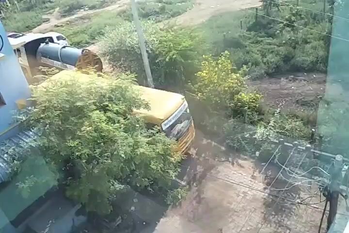 A yellow school bus is visible in the frame, partially obscured by trees. The bus appears to be in a dirt lot or road. To the left of the bus is a blue building. Further left, a white vehicle is connected to a large cylindrical tank, possibly a water tank. The environment is overgrown with vegetation, suggesting an outdoor setting. The camera angle is from above, looking down on the scene.