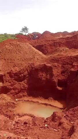 This is a short video clip showing a landscape of a construction or excavation site. The dominant color is reddish-brown earth, with piles of excavated soil and deep trenches. In the foreground, there is a pit filled with murky, greenish water. In the background, on a slight incline, a lone tree stands, and a yellow excavator is visible, suggesting ongoing work. The sky is a pale, washed-out white, indicating a bright, possibly overcast day. The overall impression is one of large-scale earthmoving activity. There is no visible text within the media. The location context provided is Barakin Columbai, Nigeria. The activity taking place is excavation, likely for construction, resource extraction, or land modification. The time of day appears to be daytime, given the bright sky. No people are visible in the clip.