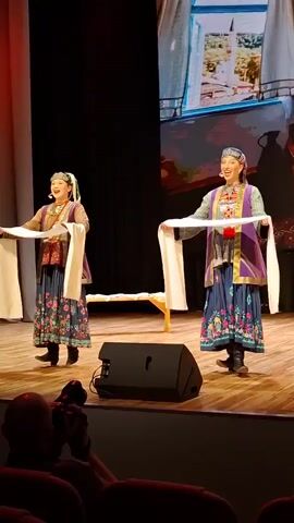 Two women are on a stage performing a dance. They are wearing traditional clothing, consisting of embroidered dresses, vests, and head coverings. Each woman is holding a long, white scarf, draped over their arms. The stage is wooden and there is a black speaker in the foreground. Behind the dancers, a large screen displays a scene of a body of water with buildings on the shore. The lighting is dim, with spotlights on the performers. The audience is not clearly visible, but parts of red seats can be seen in the foreground.