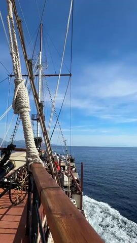 The video shows the view from the deck of a sailing ship as it moves through the water. A large rope is prominently displayed in the foreground, coiled and secured. To the left, a cannon on a wooden carriage is visible. Further back on the deck, several people can be seen, some of whom are sitting. The sea is calm and blue, with a clear sky above. In the distance, land with mountains can be seen. The ship is sailing to the right, indicated by the wake behind it. The sun is shining, casting a bright light on the scene.