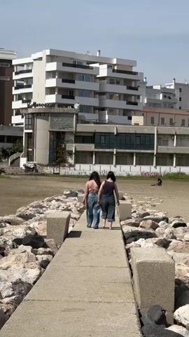Two women walk away from the camera, down a concrete pier toward a beach. They are both wearing jeans, and the woman on the left has a bare back. The pier is flanked by large rocks. A large white building is in the background. A person is seated on the beach to the right. Another person walks past on the rocks to the right. It is daytime and sunny.