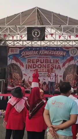 A group of people are gathered in front of a stage. On the stage, a woman in a red hijab and white dress is singing and dancing, while a man in a white shirt with a red number 17 on it stands next to her, also singing and gesturing. Behind them is a large banner that reads "SEMARAK KEMERDEKAAN REPUBLIK INDONESIA KE-81" (Independence Celebration, Republic of Indonesia, 81st Anniversary) with Indonesian flags and imagery. Several people in red shirts and hijabs are watching the performance from the audience, as are others in the background. A scaffolding structure with lights is visible above the stage. The overall environment appears to be an outdoor event.