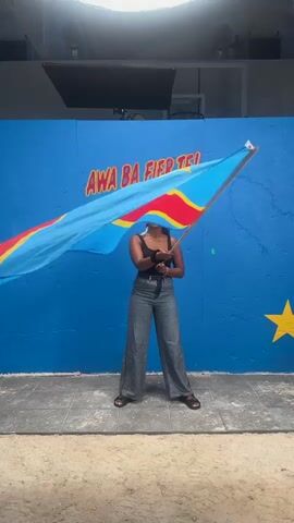 A woman stands in front of a blue wall on which is written, “AWA BA FIER TE!” in red lettering. She holds a large flag, the Democratic Republic of the Congo flag, and waves it back and forth in front of her. She is wearing a black tank top, gray pants, black sandals, and a light-colored bandana. The background is a sandy area and a gray tile floor. Overhead are various studio lights.