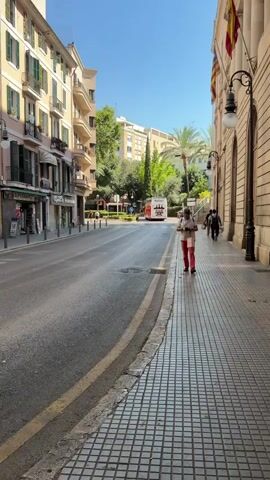 A single person walks along a sidewalk on a sunny day. The person is wearing red pants and a light-colored shirt. They are looking down at a phone in their hands. The sidewalk is paved with checkered tiles. To the left of the sidewalk is a street with a yellow line marking the edge. Across the street are buildings with shops on the ground floor. In the distance, a red tour bus is visible on the street. Trees and palm trees are also visible in the background. The sky is clear and blue.