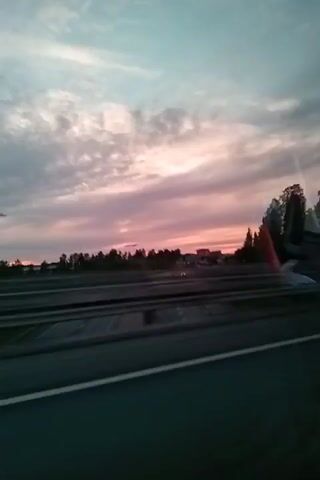 The video captures a roadside view during what appears to be sunset. The sky is a blend of soft pinks, purples, and blues, with streaks of clouds. In the foreground, a highway is visible with lane markings. To the right and slightly in the background, a line of trees is seen, their forms blurred by motion. The overall impression is one of a moving vehicle looking out at a twilight landscape.