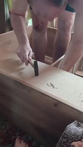 This media file captures a close-up, low-angle shot of a person assembling a wooden furniture item, possibly a cabinet or shelving unit. The person, wearing black shorts, is bent over the partially constructed furniture. Their legs and torso are visible, with some bruising on their knees and shins. They are holding a small hammer in their right hand, appearing to be in the process of hammering a nail or securing a joint. Several nails are scattered on the surface of the wooden plank they are working on, along with a measuring tape. The furniture itself is made of light-colored wood or particleboard. The background is slightly out of focus, showing fabric curtains and what appears to be a patterned rug on the floor. The lighting suggests it is daytime, but the indoor setting makes it difficult to determine the exact time or weather conditions. There are no visible text elements in the media. The overall impression is one of manual labor and DIY furniture assembly within a home environment.