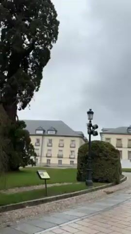 The video shows a panning view of a European-style building complex under a cloudy sky. The camera starts with a view of yellow buildings with grey roofs, a black lamp post, and a green bush. It pans to reveal a pink and grey building with two towers topped with crosses, and then pans to a beige building with a tower. The complex has a paved courtyard. The video ends with a view of the pink and grey building, the beige building, and a tall green tree.