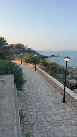 A paved walkway curves along a rocky coastline under a clear sky. The walkway is constructed from irregularly shaped stones and is bordered by a stone wall on the right. Several lampposts are spaced along the walkway, illuminating it. Lush green vegetation, including bushes and trees, grows on the hillside to the left of the walkway and beyond the wall on the right. In the distance, buildings are visible on the hillside, and the sea stretches out to the horizon. The water appears calm with some small waves breaking against the rocks. The lighting suggests it is either early morning or late afternoon.