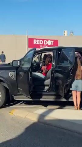 The video shows a static, medium shot of a black SUV parked outside a "RED DOT WA LOCAL" store. The driver's side door and rear passenger door are open. A person wearing a red shirt and jeans is seated in the driver's seat, looking down at their hands. Two girls stand near the open rear door; one is wearing denim shorts and a brown top, the other is wearing a light purple top and black leggings. The first girl has long brown hair, the second has long blonde hair. A man in a blue shirt walks away from the vehicle in the background. The environment appears to be a sunny day in a parking lot.
