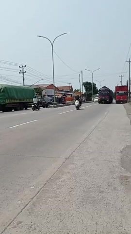 The video shows a rural road with traffic. On the left side of the road, a green truck is parked. A person on a motorcycle is riding past it. In the middle of the road, a white car is driving in the same direction as the motorcycle. Further down the road, a red truck is driving in the same direction. To the right of the road, another red truck is visible. The sky is clear and the sun is shining. There are houses and power lines visible along the side of the road.